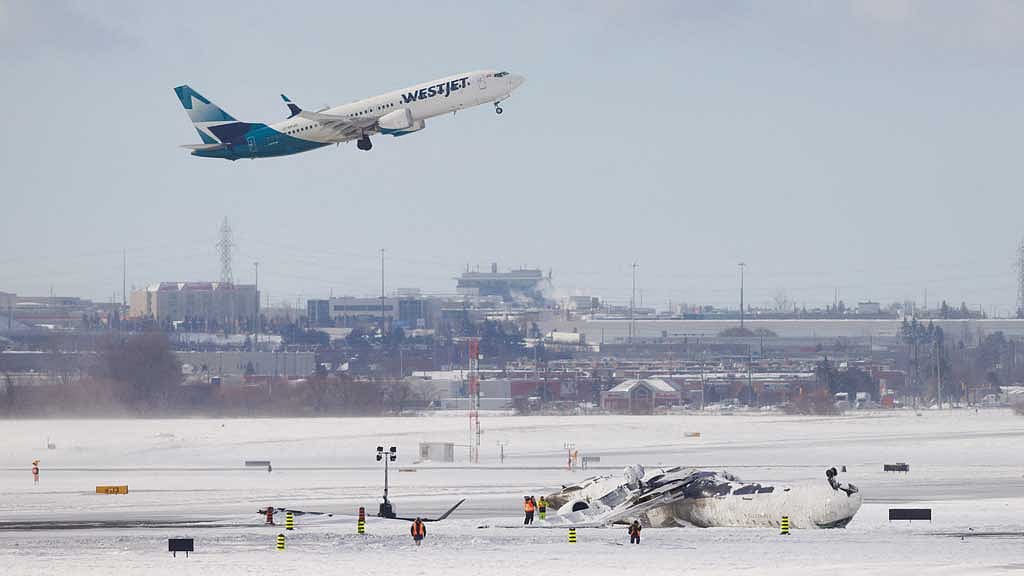 A plane takes off while a Delta Air Lines-operated CRJ900 aircraft lies on the runway following a crash at Toronto Pearson International Airport in Mississauga, Ontario, Canada Feb 18, 2025. REUTERS/Cole Burston